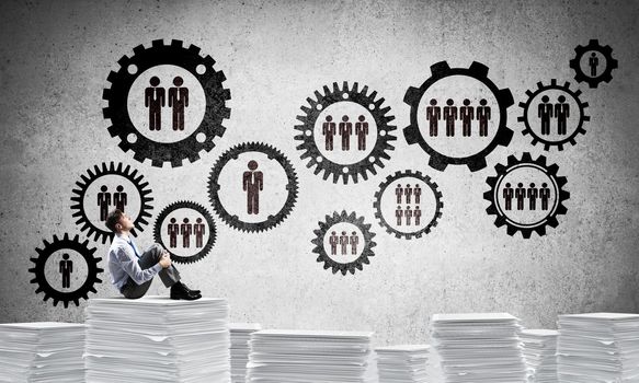 Businessman looking away while sitting on pile of documents against social gear structure on background. Mixed media.