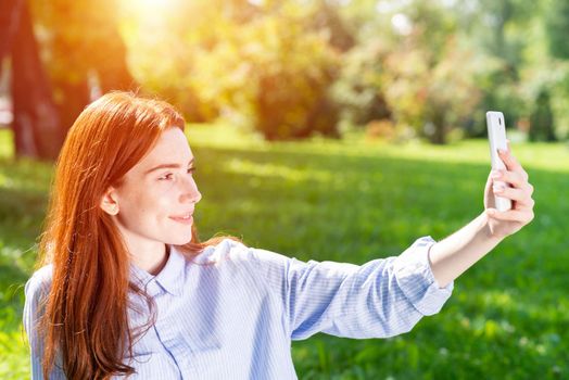 Young redhead woman taking selfie photo on green field. Close up portrait of charming girl with smartphone in hand. Lady having fun in spring park at sunny summer day. Digital mobile technology