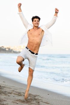 Portrait of an attractive young man jumping on a tropical beach. Handsome guy wearing white shirt and striped shorts. Male with beautiful body.