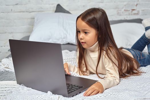 Cute little girl girl feeling amusing while watching cartoons on a laptop sitting on bed. White sweater and jeans.