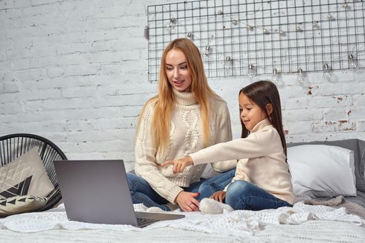 Beautiful young mother and her cute daughter in white sweaters and jeans lying on the bed at home, laughing and looking in laptop. Family time