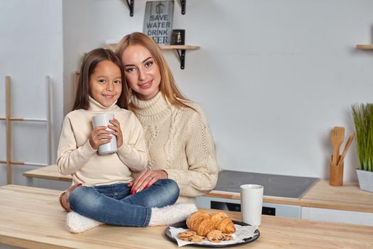 Shot of cheerful mother and daughter sit together at kitchen table, drink hot tea in morning, have pleasant friendly talk between each other. Curious girl asks something in mum during coffee break.