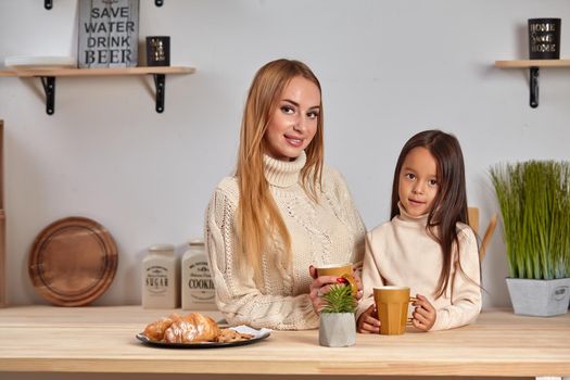 Shot of cheerful mother and daughter sit together at kitchen table, drink hot tea in morning, have pleasant friendly talk between each other. Curious girl asks something in mum during coffee break.