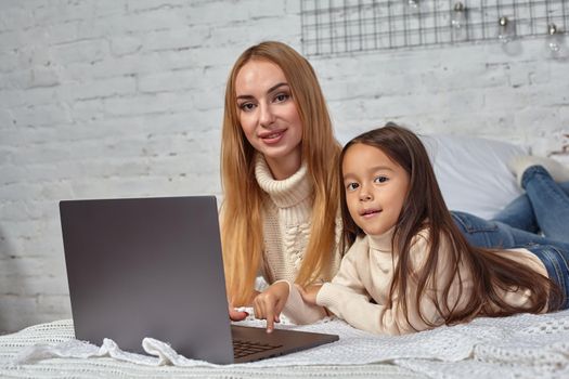 Beautiful young mother and her cute daughter in white sweaters and jeans lying on the bed at home, laughing and looking in laptop. Family time