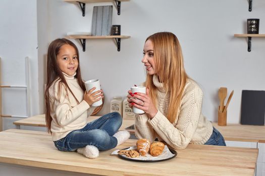 Shot of cheerful mother and daughter sit together at kitchen table, drink hot tea in morning, have pleasant friendly talk between each other. Curious girl asks something in mum during coffee break.