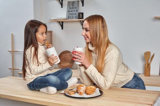 Shot of cheerful mother and daughter sit together at kitchen table, drink hot tea in morning, have pleasant friendly talk between each other. Curious girl asks something in mum during coffee break.