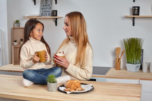 Shot of cheerful mother and daughter sit together at kitchen table, drink hot tea in morning, have pleasant friendly talk between each other. Curious girl asks something in mum during coffee break.