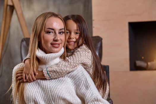 Beautiful mother woman in white sweater hugs her little daughter. Cheerful playful mood and love inside the family. Cozy home environment.