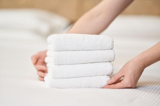 Close up of female hands putting stack of fresh white bath towels on the bed sheet in room. Hotel service concept
