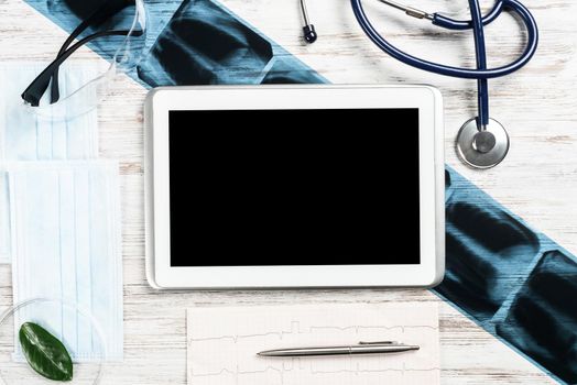 Flat lay workplace of doctor in modern clinic. Tablet computer with blank screen, x-ray image, stethoscope and cardiogram lies on wooden desk. Professional medical consultation and treatment