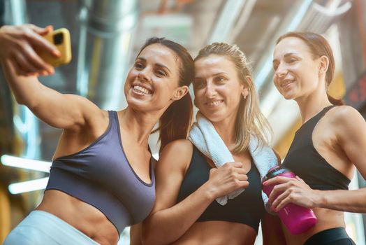 Happy moments. Group of three young beautiful and cheerful sportive women taking selfie on smartphone while resting after workout, exercising together at gym. Sport, wellness and healthy lifestyle