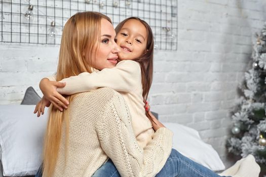 Beautiful young woman and her charming little daughter in white sweaters and jeans lying on the bed at home, are hugging.