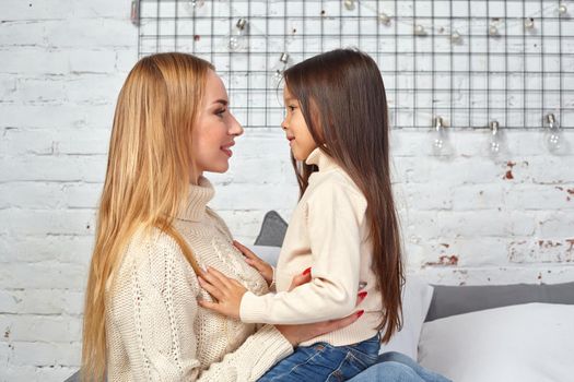 Beautiful young woman and her charming little daughter in white sweaters and jeans lying on the bed at home, are hugging.