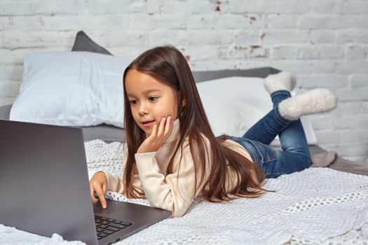 Cute little girl girl feeling amusing while watching cartoons on a laptop sitting on bed. White sweater and jeans.