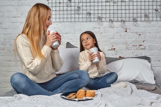 Mother and daughter in white sweaters and jeans at home, drink tea or cocoa from large mugs sitting on the bed