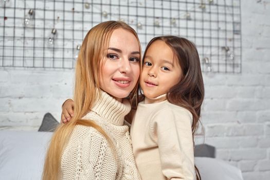 Beautiful young woman and her charming little daughter in white sweaters and jeans lying on the bed at home, are hugging.