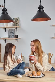 Shot of cheerful mother and daughter sit together at kitchen table, drink hot tea in morning, have pleasant friendly talk between each other. Curious girl asks something in mum during coffee break.