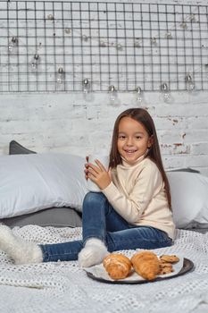 Pretty little girl 6 years old in a white sweater and jeans. Child in the room with a bed, eating croissant and drinking tea or cocoa, good mood.