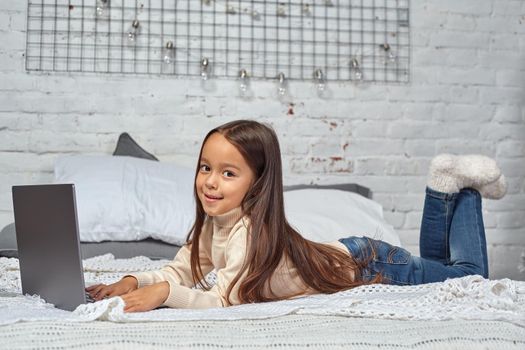 Cute little girl girl feeling amusing while watching cartoons on a laptop sitting on bed. White sweater and jeans.