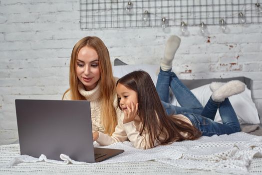 Beautiful young mother and her cute daughter in white sweaters and jeans lying on the bed at home, laughing and looking in laptop. Family time