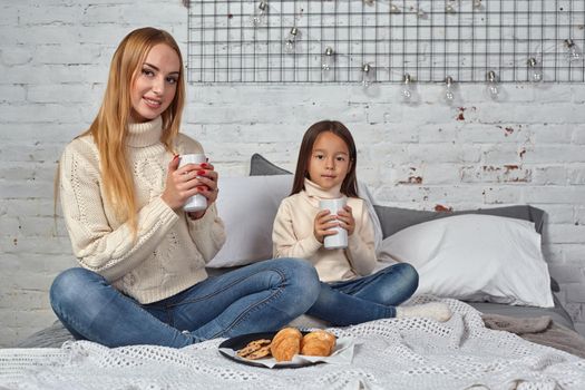 Mother and daughter in white sweaters and jeans at home, drink tea or cocoa from large mugs sitting on the bed