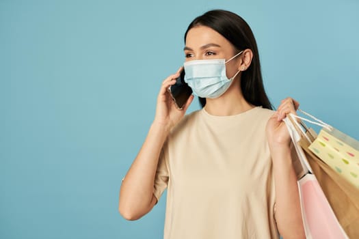 Waist up of pretty lady in disposable face mask holding shopping bags while talking on mobile phone. Copy space. Quarantine, coronavirus concept