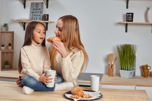 Shot of cheerful mother and daughter sit together at kitchen table, drink hot tea in morning, have pleasant friendly talk between each other. Curious girl asks something in mum during coffee break.