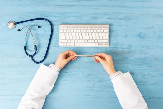 Top view of doctor hands holding pen. Physician in medical uniform working at blue wooden desk. Examination and diagnosis in hospital. Practitioner workplace with stethoscope and computer keyboard.