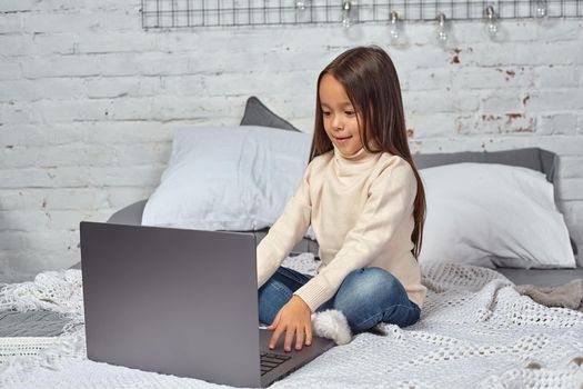 Cute little girl girl feeling amusing while watching cartoons on a laptop sitting on bed. White sweater and jeans.