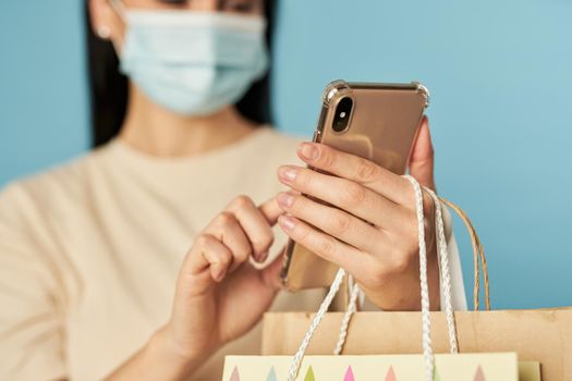 Waist up of pretty female in disposable face mask holding shopping bags while typing a message on the phone. Copy space. Quarantine, coronavirus concept