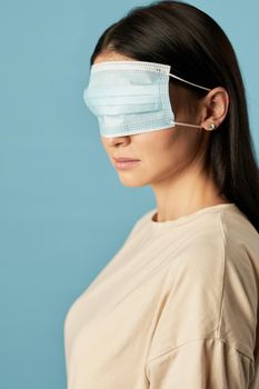 Side view of woman in a beige t-shirt posing with a protective mask on her eyes on a blue background. Copy space. Quarantine, epidemic concept