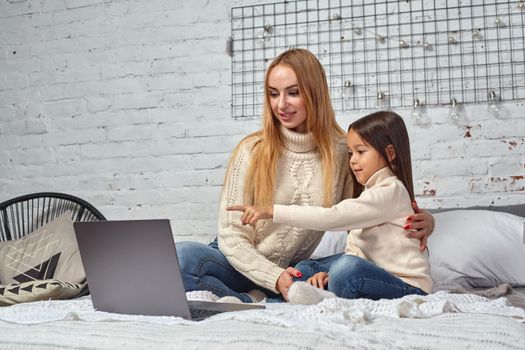 Beautiful young mother and her cute daughter in white sweaters and jeans lying on the bed at home, laughing and looking in laptop. Family time