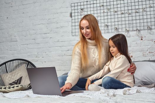 Beautiful young mother and her cute daughter in white sweaters and jeans lying on the bed at home, laughing and looking in laptop. Family time