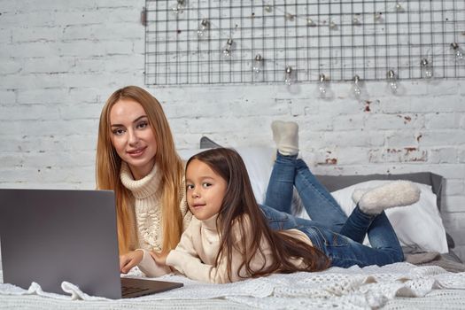 Beautiful young mother and her cute daughter in white sweaters and jeans lying on the bed at home, laughing and looking in laptop. Family time