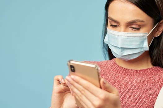 Cropped photo of pretty young woman wearing protective mask and typing message on mobile phone, isolated on blue background. Copy space. Quarantine, coronavirus concept