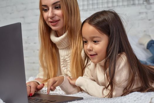 Beautiful young mother and her cute daughter in white sweaters and jeans lying on the bed at home, laughing and looking in laptop. Family time