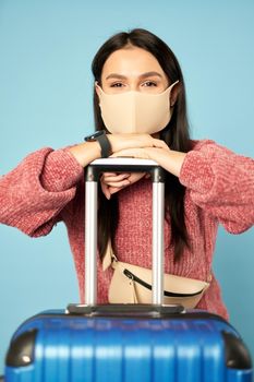 Young female sitting next to suitcase while wearing protective mask posing in studio. Copy space. Concept of travel, coronavirus