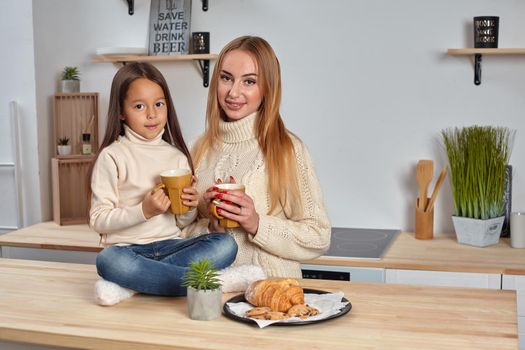 Shot of cheerful mother and daughter sit together at kitchen table, drink hot tea in morning, have pleasant friendly talk between each other. Curious girl asks something in mum during coffee break.