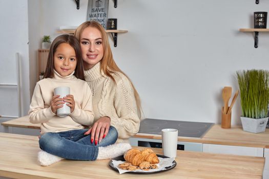 Shot of cheerful mother and daughter sit together at kitchen table, drink hot tea in morning, have pleasant friendly talk between each other. Curious girl asks something in mum during coffee break.