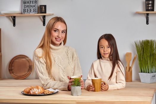 Shot of cheerful mother and daughter sit together at kitchen table, drink hot tea in morning, have pleasant friendly talk between each other. Curious girl asks something in mum during coffee break.