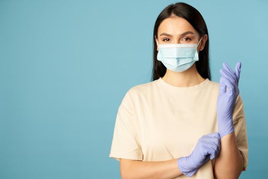 Waist up of pretty woman in a beige t-shirt standing in the studio in a protective mask and gloves on a blue background. Copy space. Quarantine, epidemic concept