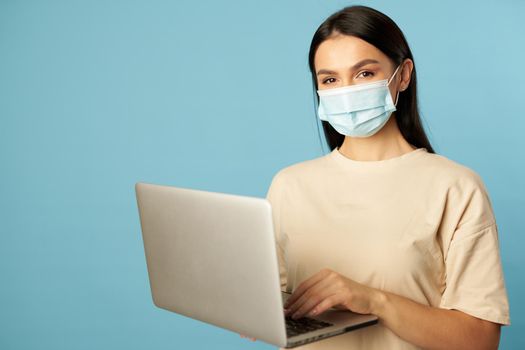 Young woman in beige t-shirt wearing protective mask and holding laptop in the studio on blue background. Copy space. Quarantine, epidemic concept