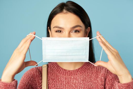 Portrait of beautiful woman posing in studio and holding face mask, isolated on blue background. Quarantine, coronavirus concept