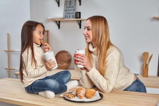 Shot of cheerful mother and daughter sit together at kitchen table, drink hot tea in morning, have pleasant friendly talk between each other. Curious girl asks something in mum during coffee break.