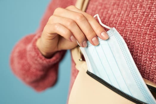 Close up of female holding protective face mask and taking out of her bag on a blue background. Copy space. Quarantine, coronavirus concept