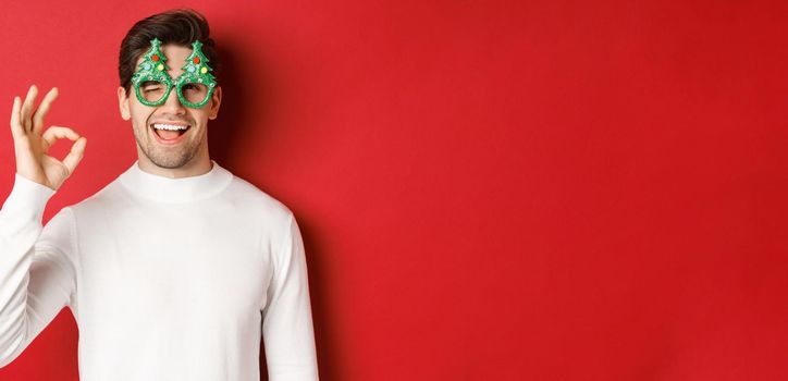 Portrait of handsome and cheeky guy, wearing party glasses and white sweater, showing okay sign and winking, wishing happy new year, standing over red background.