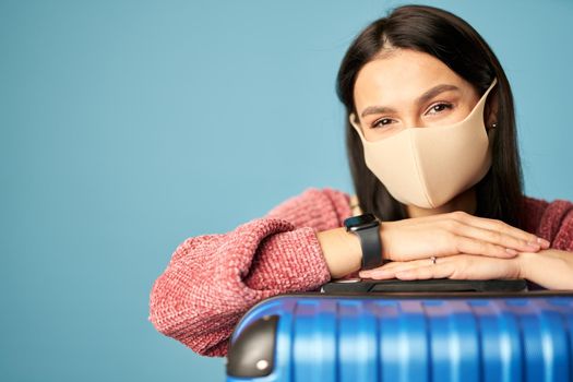 Caucasian female tourist wearing an anti-virus mask with suitcase posing against blue background. Copy space. Concept of travel, coronavirus