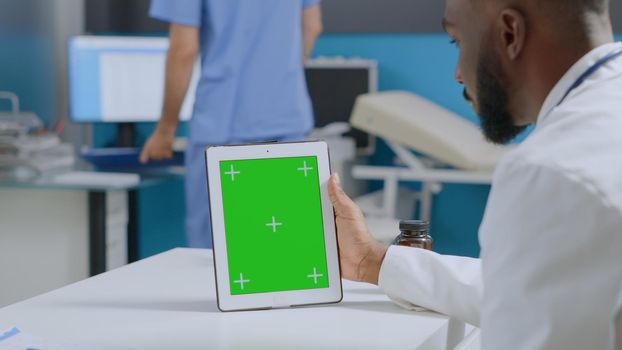 African american specialist doctor looking at mock up green screen chroma key tablet computer with isolated display. Therapist man working at healthcare treatment in hospital office. Medicine services