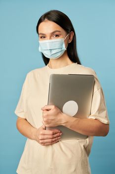 Portrait of pretty lady posing in studio while wearing mask and using laptop against blue background. Copy space. Quarantine, epidemic concept