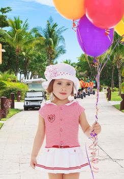 Adorable little girl in short summer dress and white Panama hat on his head. Girl holding bunch of colorful balloons.Close-up.On the background of the track